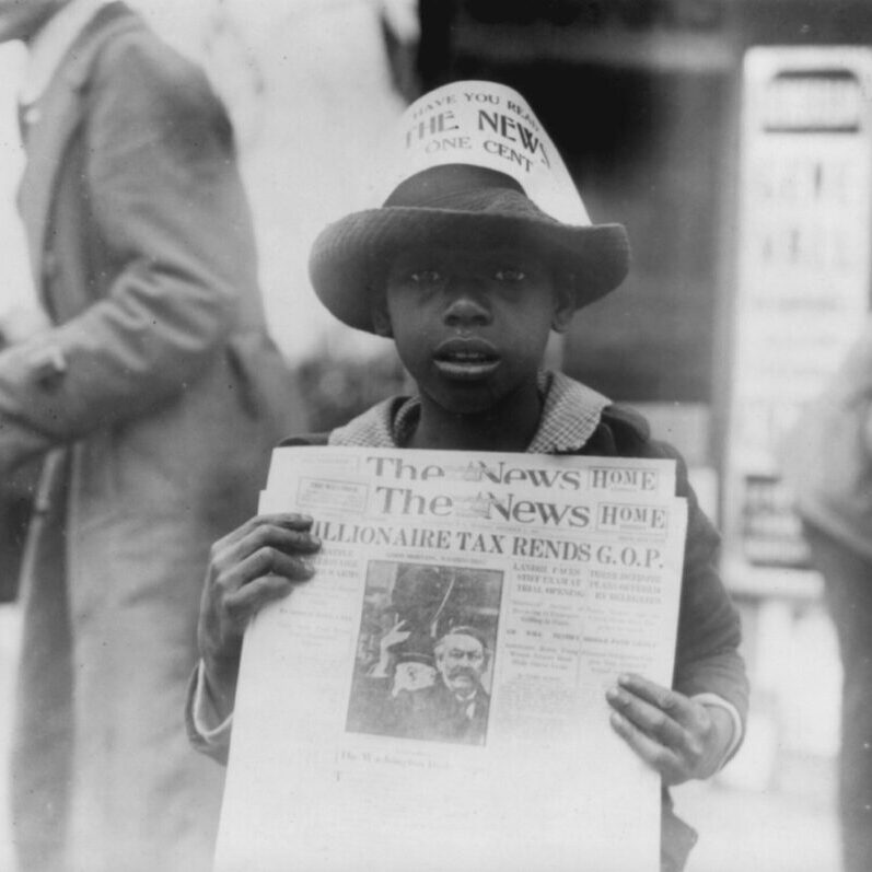 A young boy holding up a newspaper in front of him.